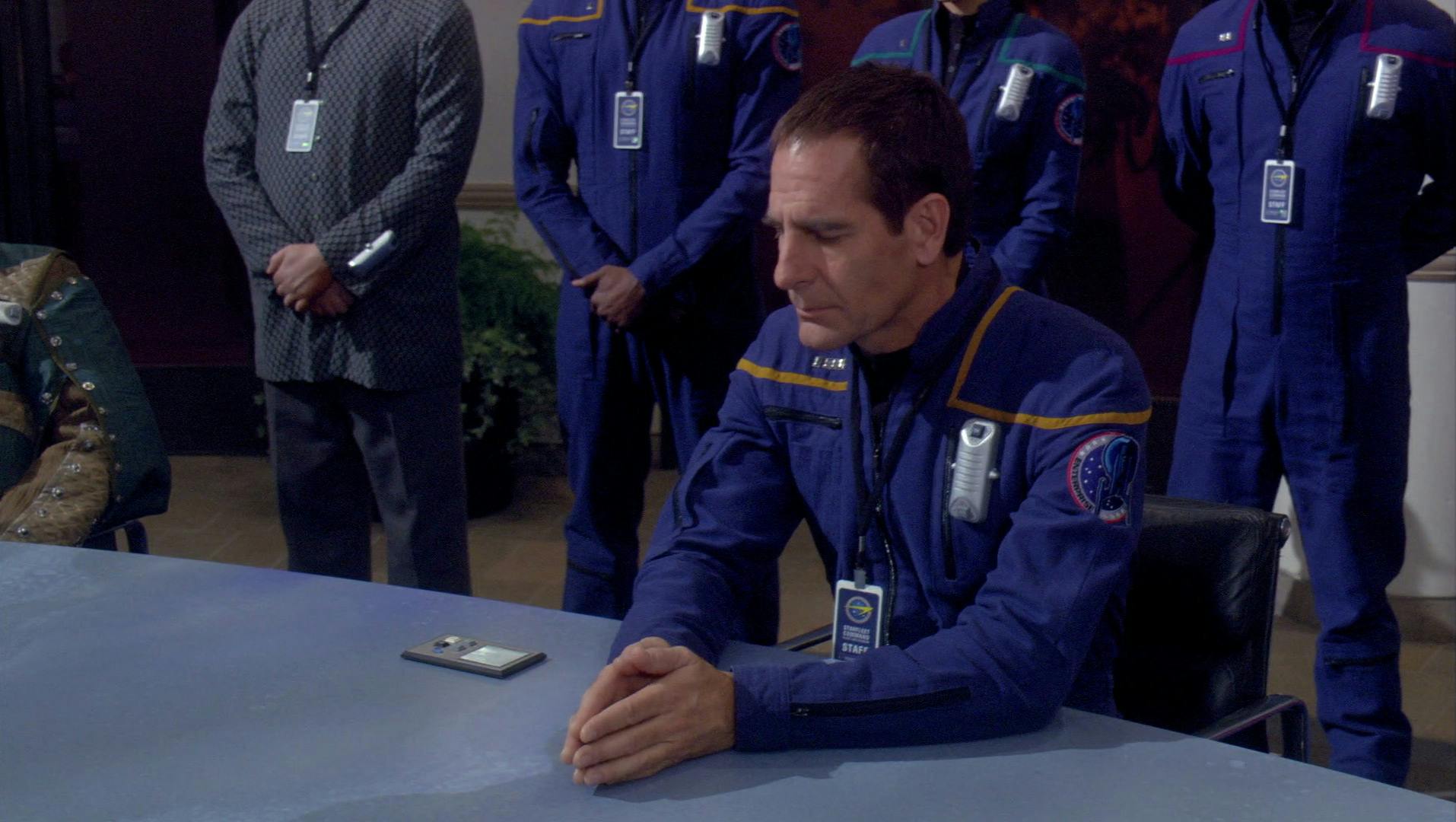 Archer solemnly sits with the palm of his hands touching with his crew standing behind him in 'Terra Prime'