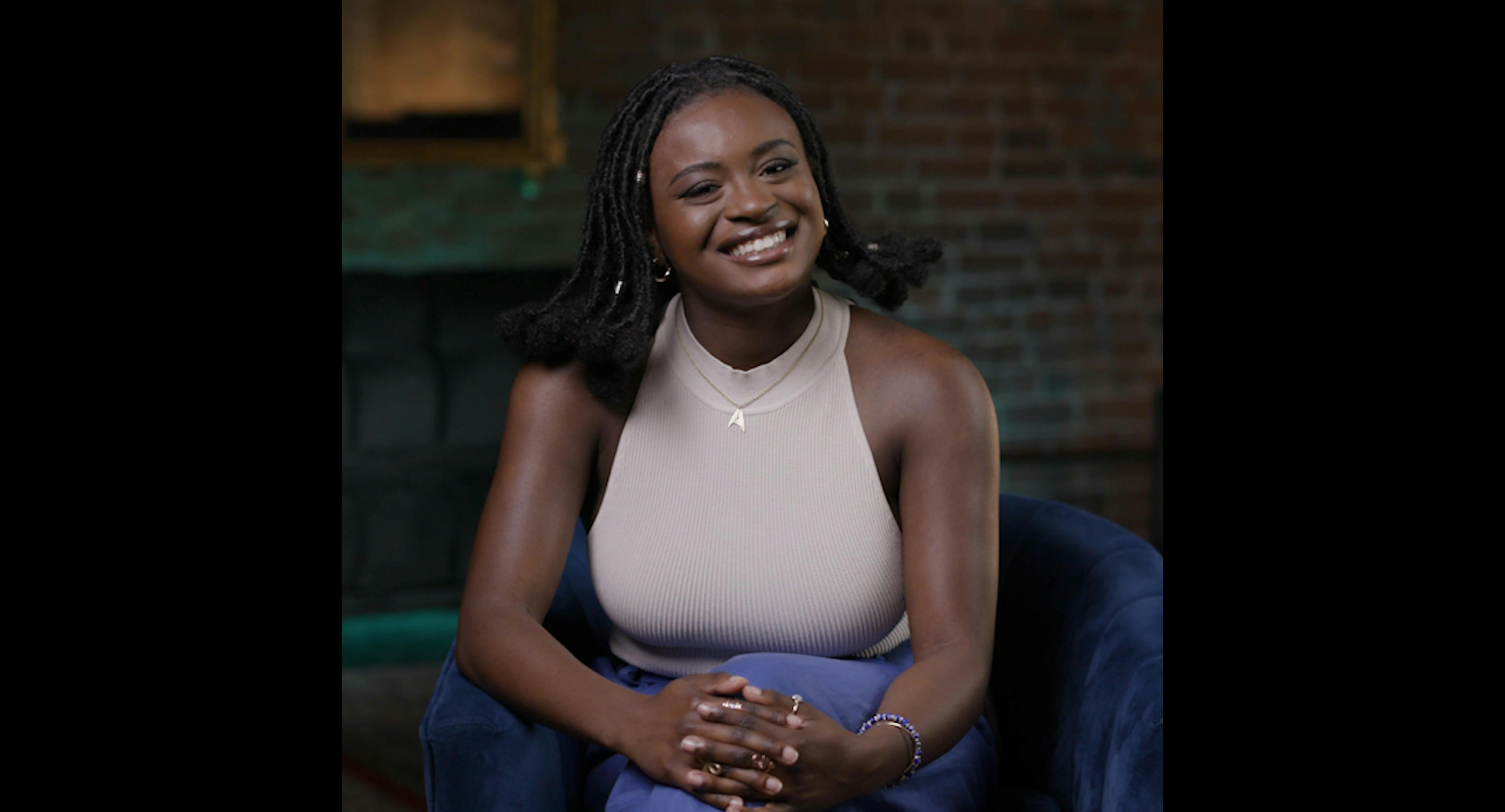 Celia Rose Gooding, with her legs crossed and hands folded on her knee, smiles while looking straight ahead