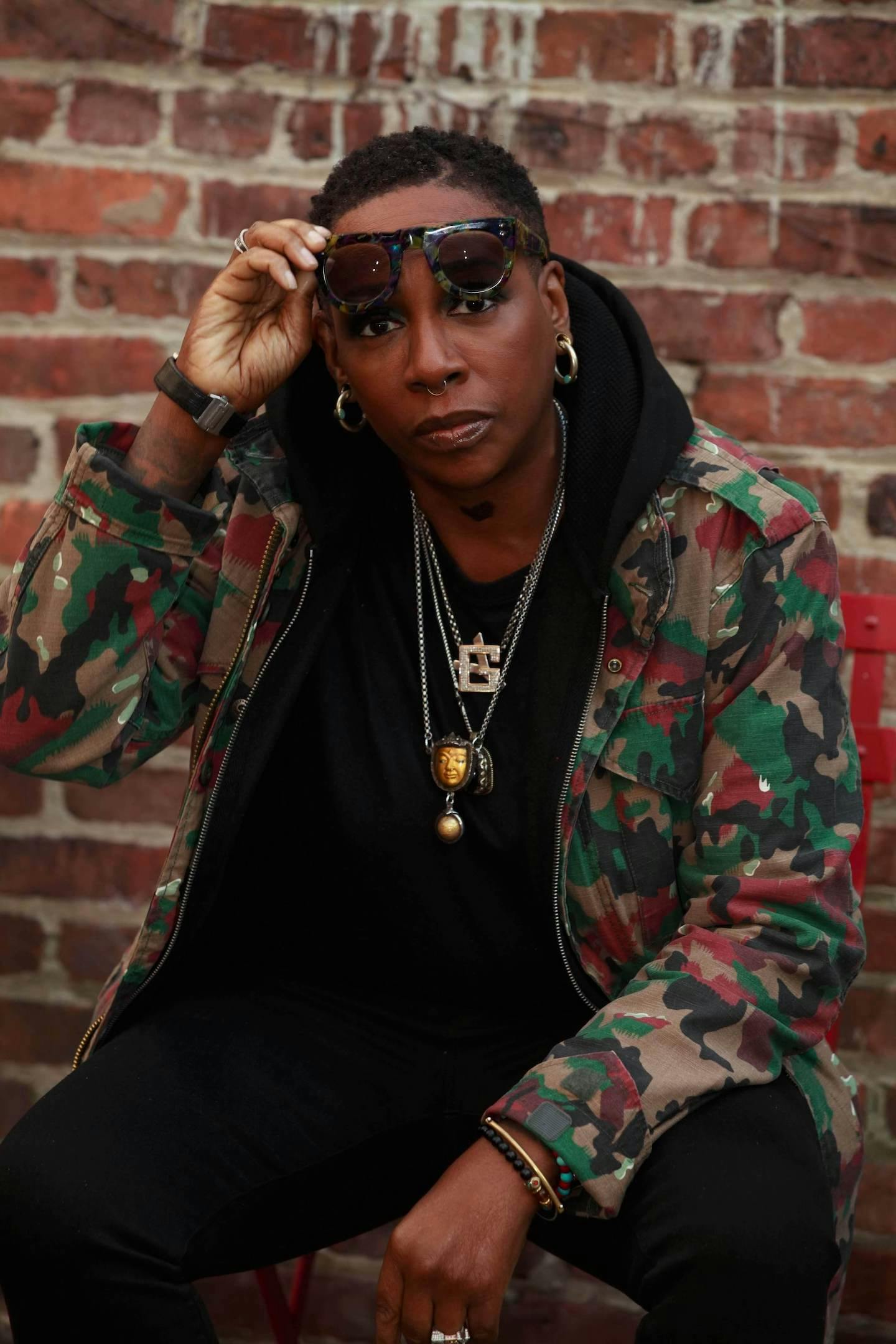 Professional headshot of Gina Yashere seated against a brick wall raising sunglasses